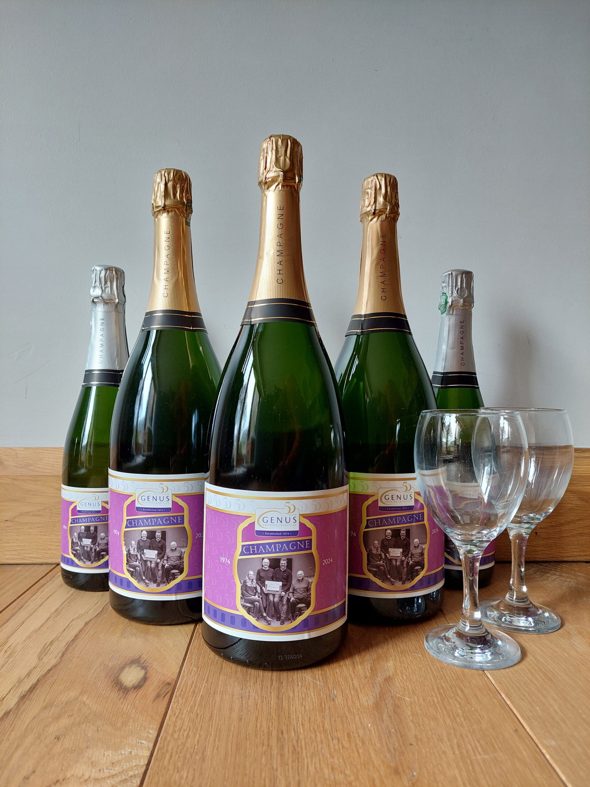 Three Genus branded Magnums of Champagne and two smaller bottles, plus two glasses displayed in a triangular formation on a wooden floor.